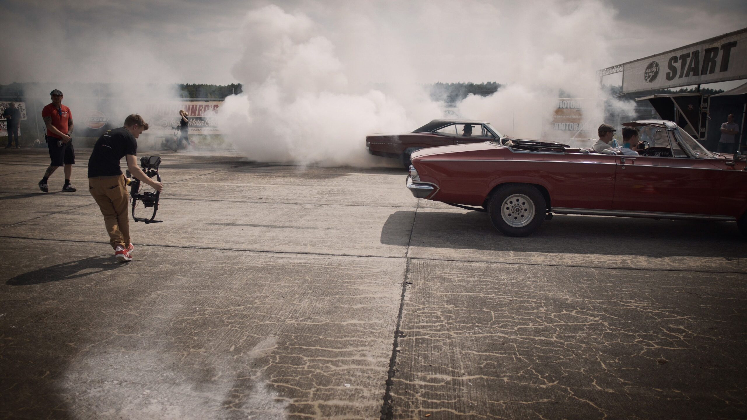 A dynamic scene of classic cars and filmmakers capturing a smoky drag race event.
